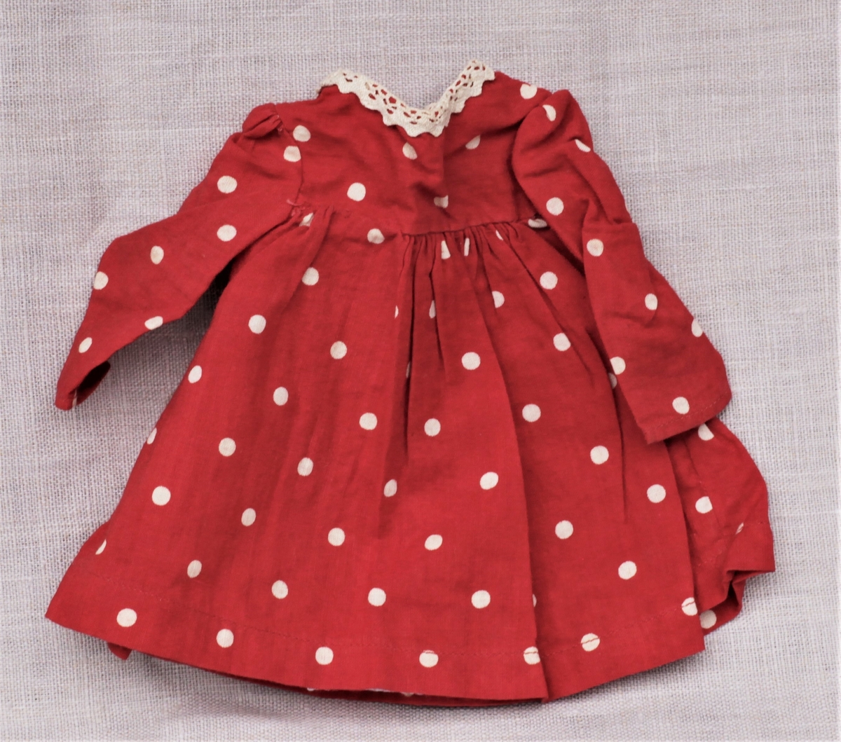 Tre enkle dukkekjoler, to rødprikkete (a og b) og en blårutet (c), en stråhatt (d), et bånd (e). Rød kjole (b) uten ermer, med hvite kanter.