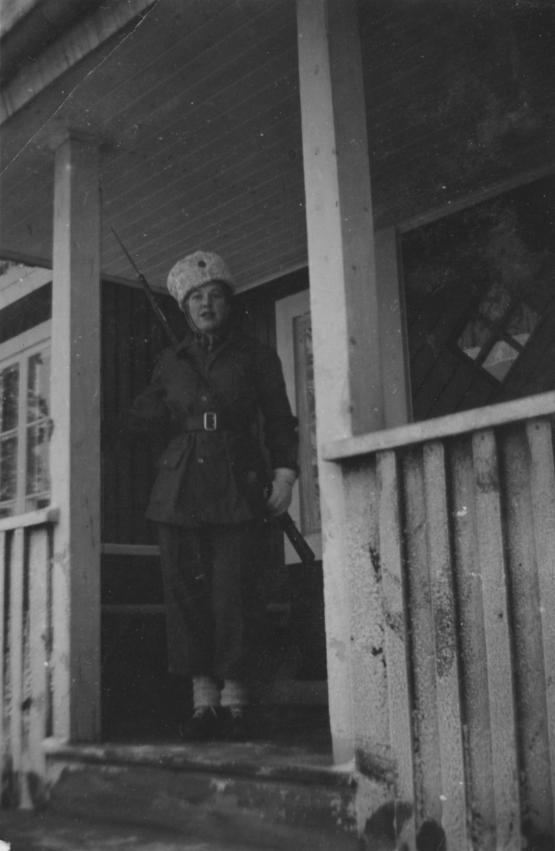 Porträtt av luftbevakare Karin Nordberg i uniform och med gevär, vintern 1942. Utomhus på verandan till luftbevakningsstationen för 91:a ls-kompaniet i Tellejåkk, Kåbdalis under beredskapsåren.