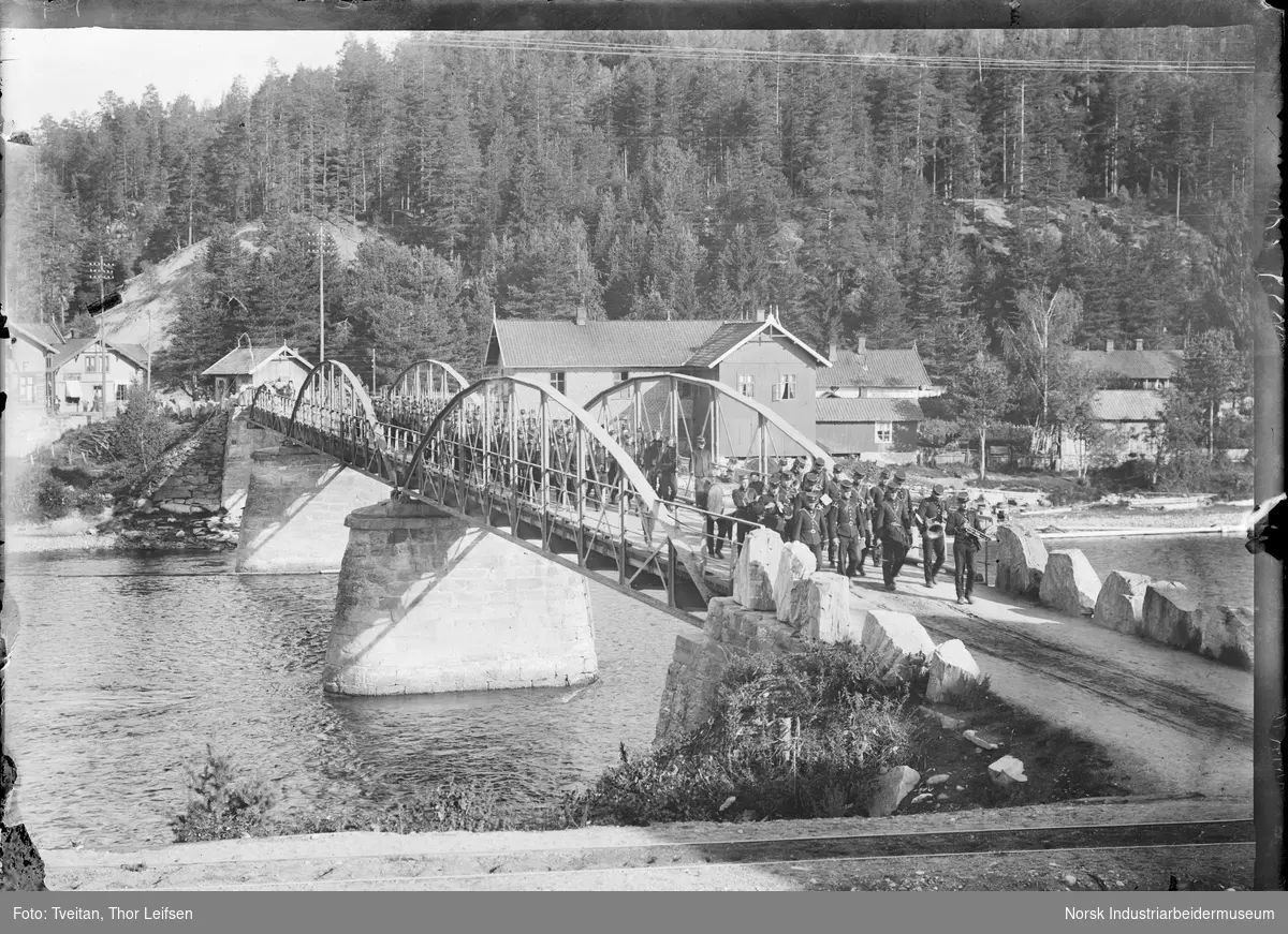 Menn i uniform og noen med musikkinstrument marsjerende over Tinnå bro ved nedre bydel i Notodden.