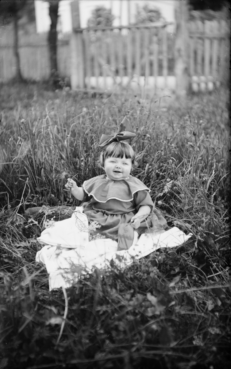 Lita jente med som sitter på et teppe i gresset, med sløyfe i håret og påsatt krave på kjolen.