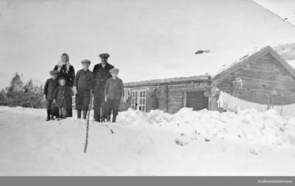 Bildet er tatt på gårdsplassen på Kvamsmoen, gruppe på seks personer hvorav fire barn, utenfor en mindre bygning, klær henger på snora?, det er vinter.