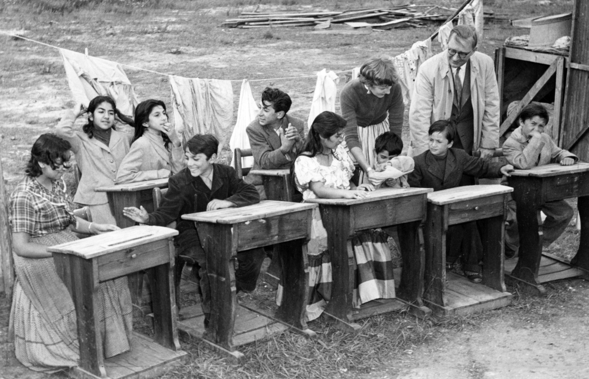 Skolundervisning för romska barn vid Frihamnen i Malmö 1954. Rektorn är på besök. I bakgrunden hänger kläder på tork i lägret.