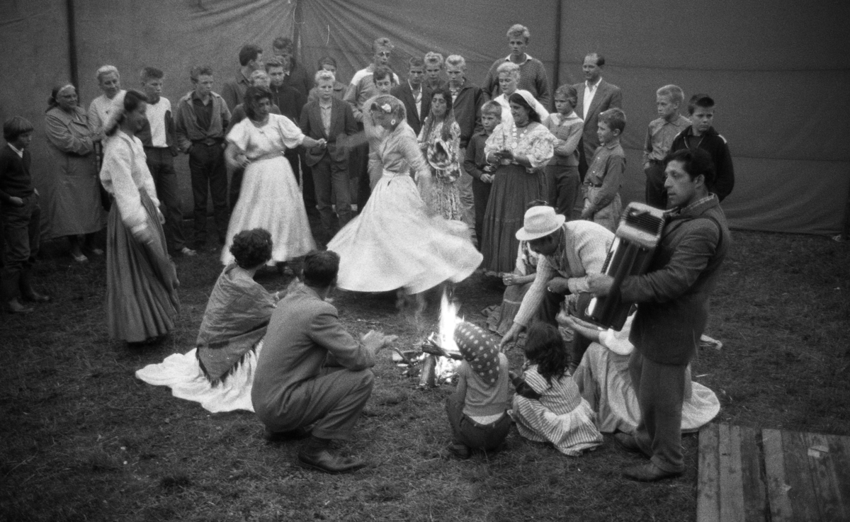 En grupp romer dansar och spelar musik kring en brasa. I bakgrunden syns tältdukar som skärmar av lägret.