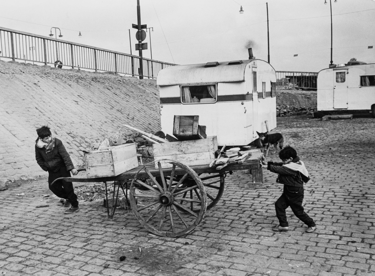 Två romska barn forslar trälådor på en kärra för att kunna göra en eld. I bakgrunden syns husvagnar.
