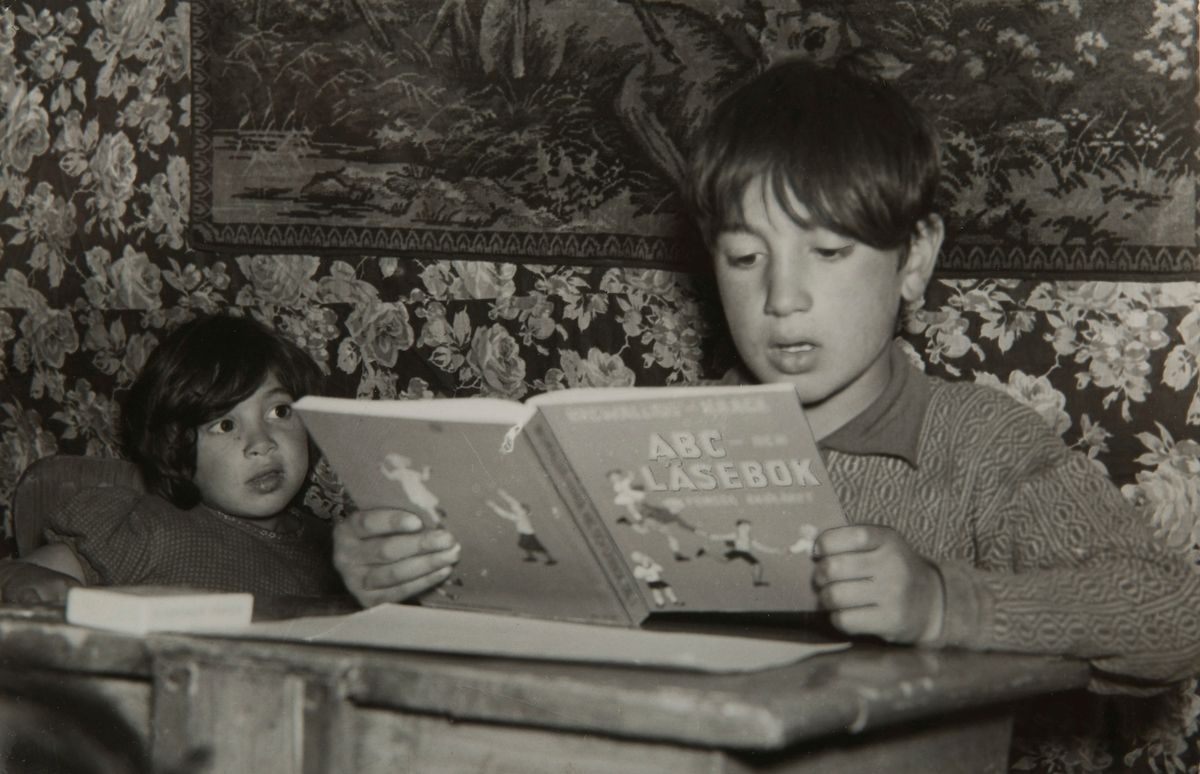 En liten pojke läser ur sin läsebok, hans lillasyster tittar på. Pojken går i sommarskola i lägret där han bor.