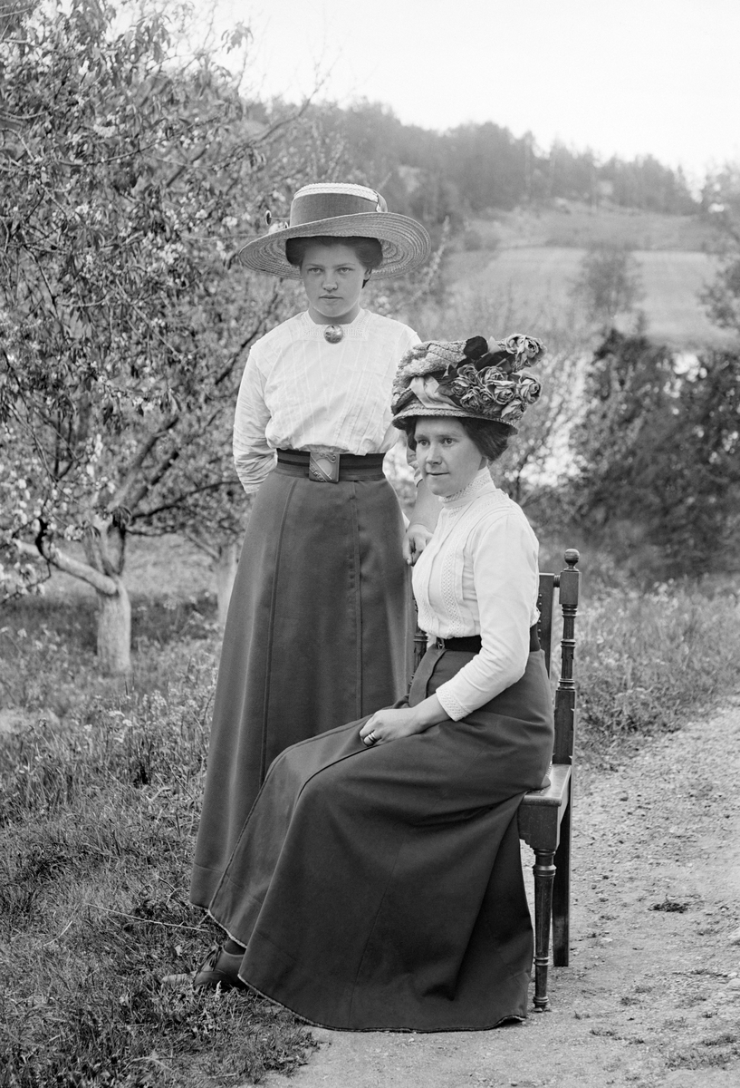 Två väl skrudade kvinnor poserar för fotograf Emil Durling vid Strömmen i Sankt Anna. Kvinnan till vänster är Magda Svensson, vidare gift med handelsbiträdet Mats Olivio Axelsson. Kvinnan på stolen är möjligtvis Magdas mor, men antagandet har inte kunnat styrkas.