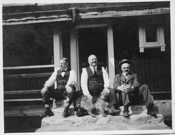 Tre menn og en hund sitter på trappa til et hus.