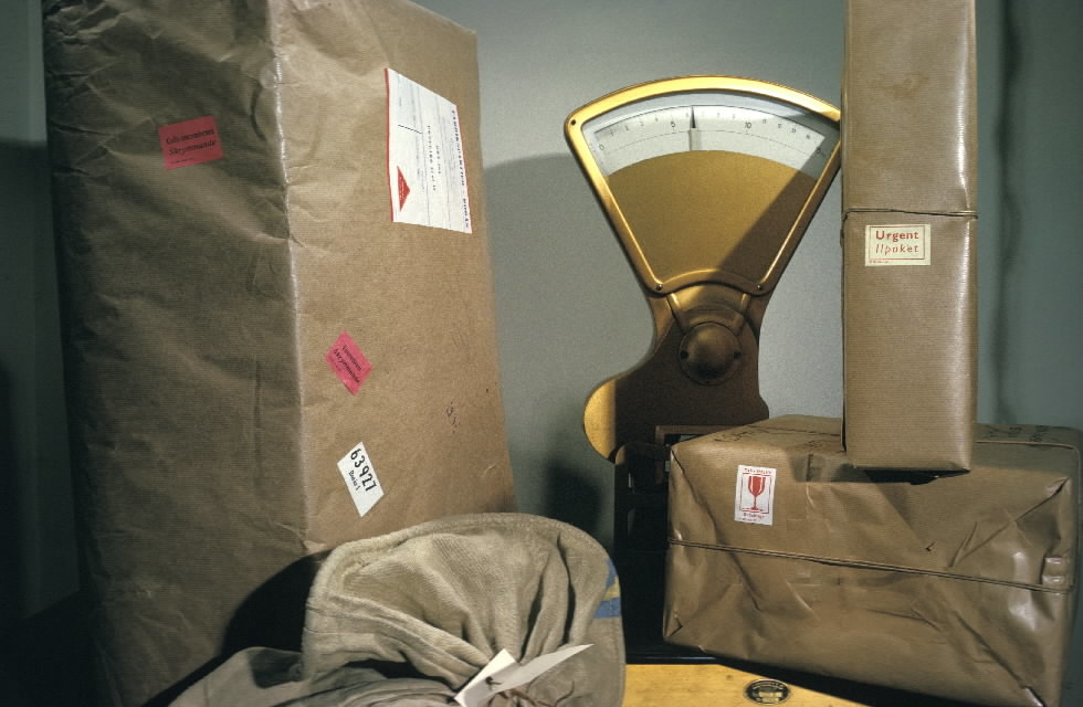 Seriebild H 13a. Paketvåg omgiven av paket med olika etiketter: 
"Skrymmande", "Bräckligt" och "Ilpaket". På postkontoret Farsta 1.