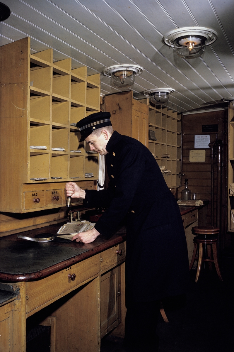I en ålderdomlig järnvägspostkupé, i Järnvägsmuseum i Tomteboda, Solna, stämplar en postiljon avgående post. I den arrangerade situationen agerar fotobiträdet Gunnar Svensson, Postens Fotografer, i en uniform från Postmuseum.