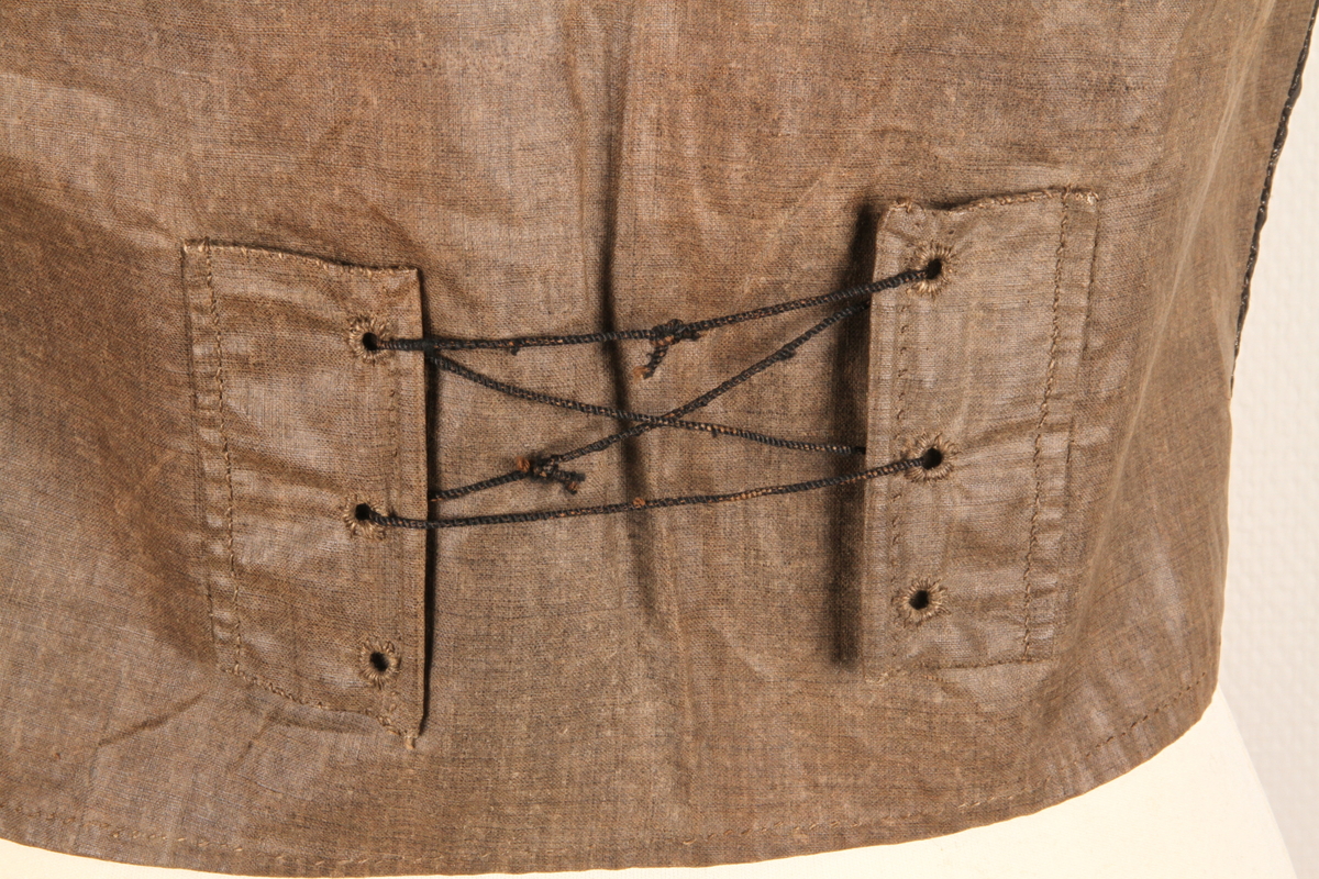 Enkeltkneppet silkevest i svart silkebrokade. Silkebrokaden har innvevde sølvtråder som danner mønsteret. Rygge er i brunt linstoff, og har snøring midt bak. Vesten er foret med hvitt linstoff. Hele vesten er sydd for hånd, i hovedsak med lintråd. 