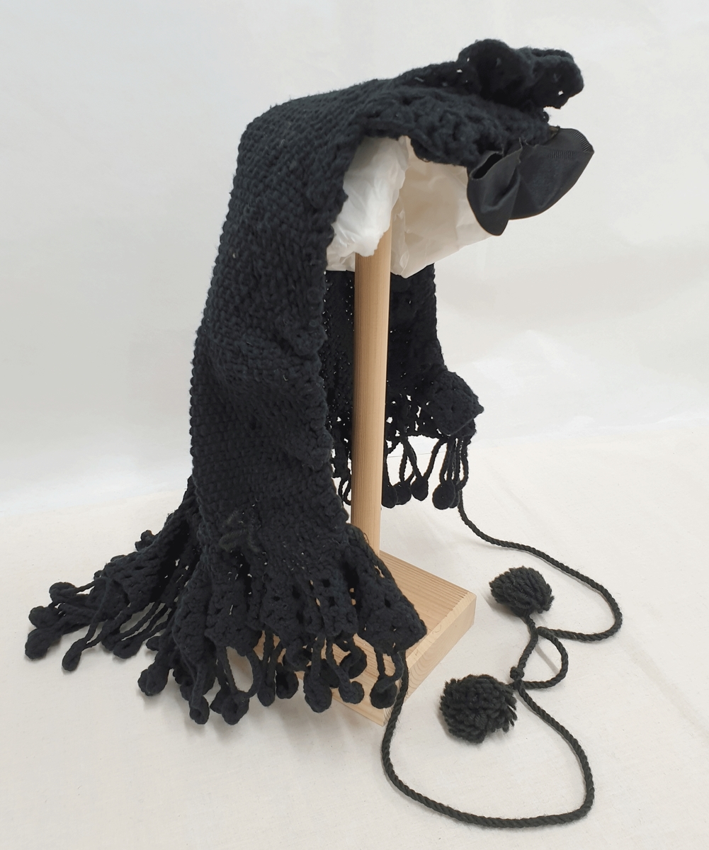 svart, strikket hodeplagg med silkesløyfe midt foran. Kantene har strikkede "blonder" og dusker langs kanten. Lange snorer med dusker.