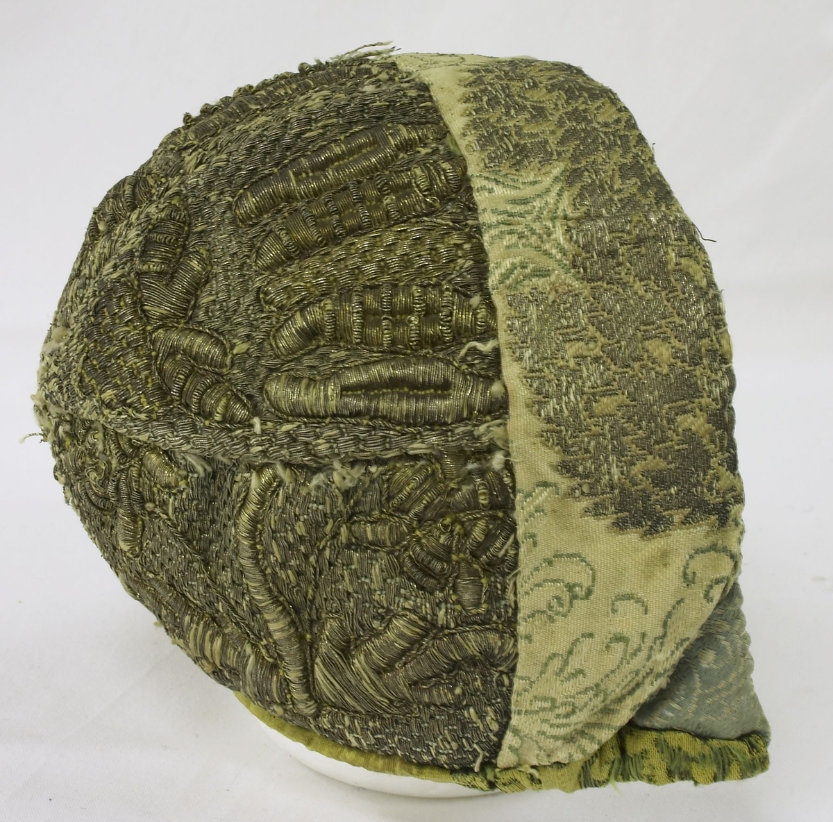 Barnelue / dåpslue. Hele lua er metallbrodert. Gul silkebrokade med mønster i grønt og svart. Fór i trykt bomull. Lua er vatert. Hele lua er sydd for hånd. Kan være fra 1700-tallet.