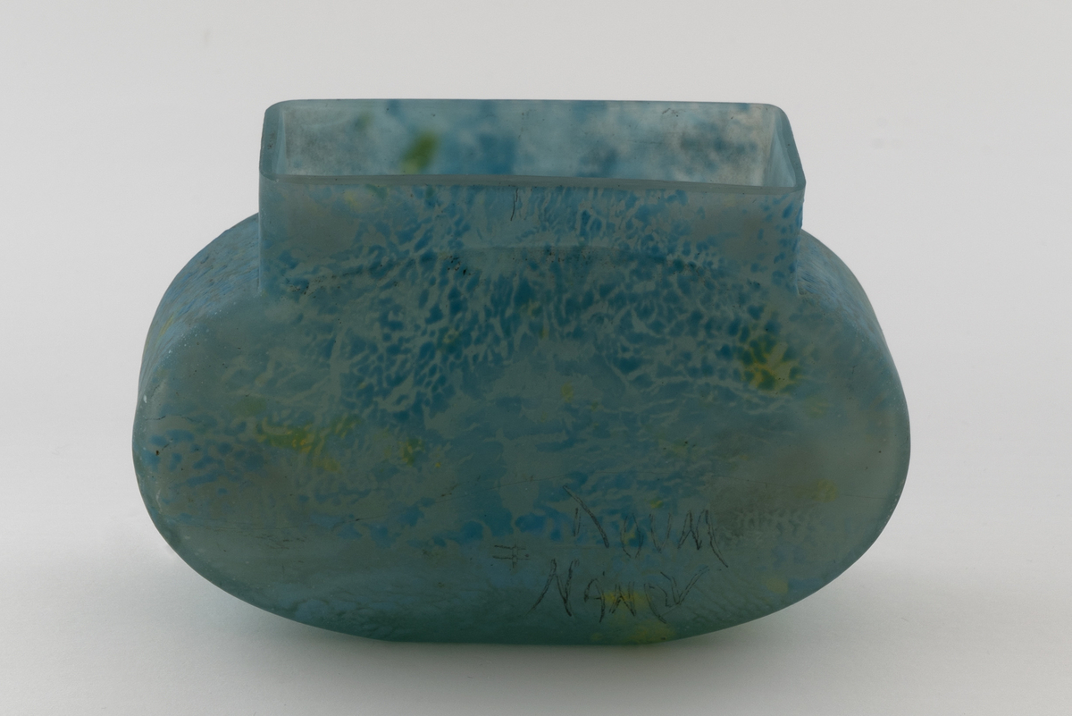 Vase utført i halvgjennomskinnelig overfangsglass. Korpus er ovalformet og lett utbuet. Åpningen er rektangulær med forhøyede kanter. Vasen har en kjerne i klart glass. Yttersiden av korpus er dekket av et blått sjikt, og er dekorert med partier i blått, gult og grønt som danner et marmoreringslignede mønster på vasens overflate.