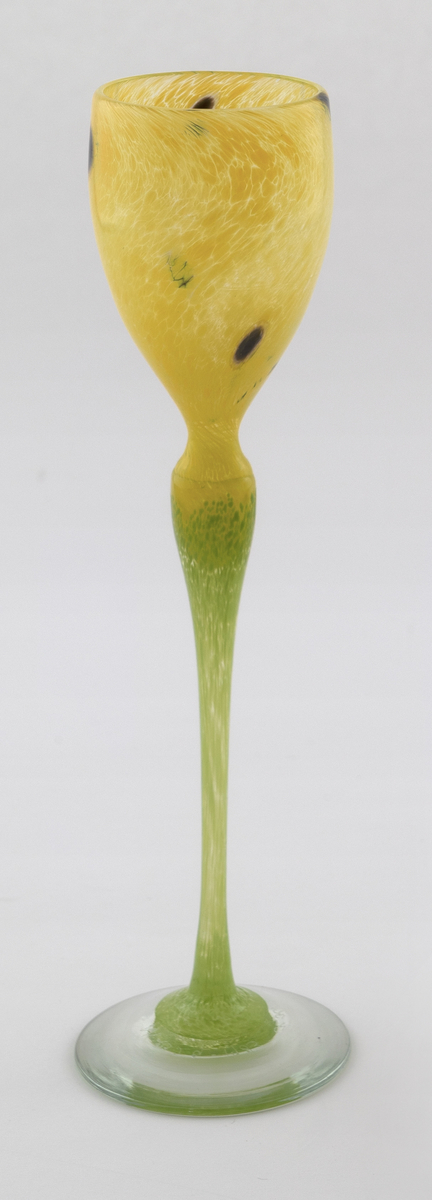 Høyt drikkeglass med tulipanlignede utforming. Eggeformet kupa i gult halvgjennomskinnelig glass med sorte prikker. Stetten er utført i lysegrønt opakt glass, som hviler på en transparent fot.