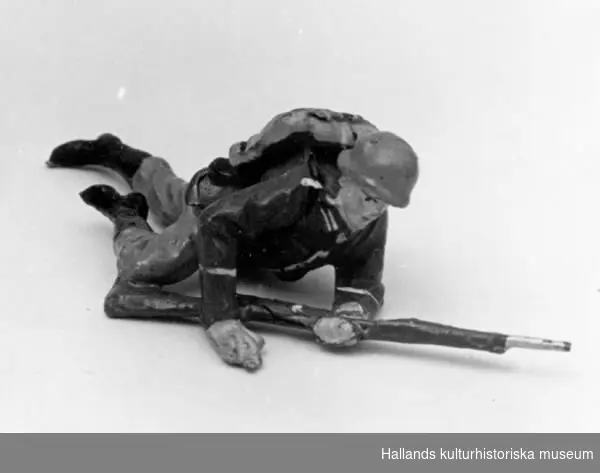 Leksakssoldat av trämassa på ståltrådsskelett i tysk uniform. Soldaten ligger ner och ålar sig fram. Målad i grönt, brunt, svart och skärt.