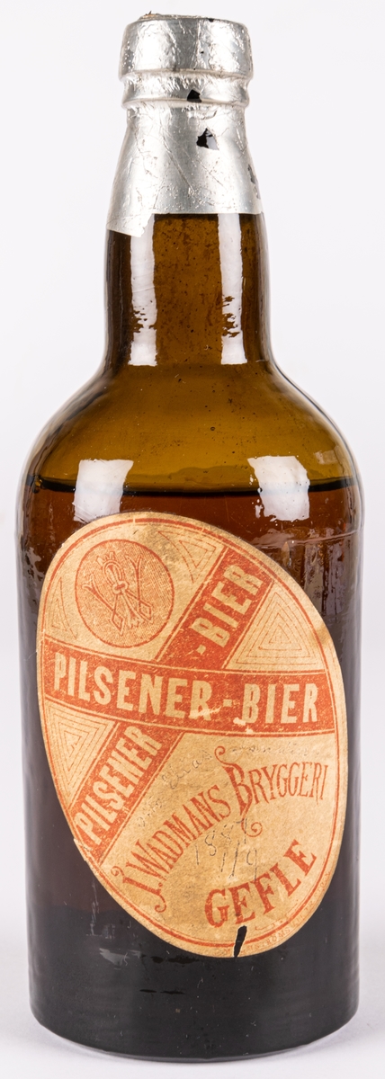 Pilsnerflaska med innehåll, enligt etiketter: Pilsener-Bier. Från J. Wadmans bryggeri, Gefle.
Brunt glas, folie över kork. Etikett av papper.