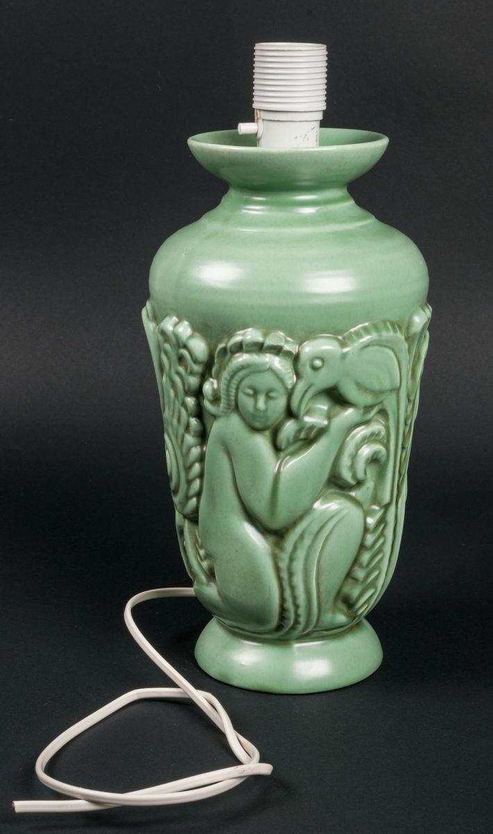Lampfot i keramik, grön glasyr 2853, med vit elektiskt sladd och lamphållare. Ingen skärm. Formpressat motiv i form av människofigurer och fåglar.