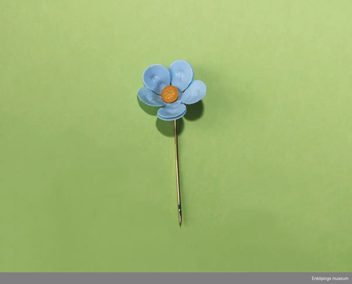 Majblomma från år 1932.
Blomman är gjord av ljusblå celluloid och har fem blad i dubbla lager, totalt 10 blad och en gul mittknapp, även denna gjord av celluloid. 
Det som håller blomman samman är en nål av mässing.