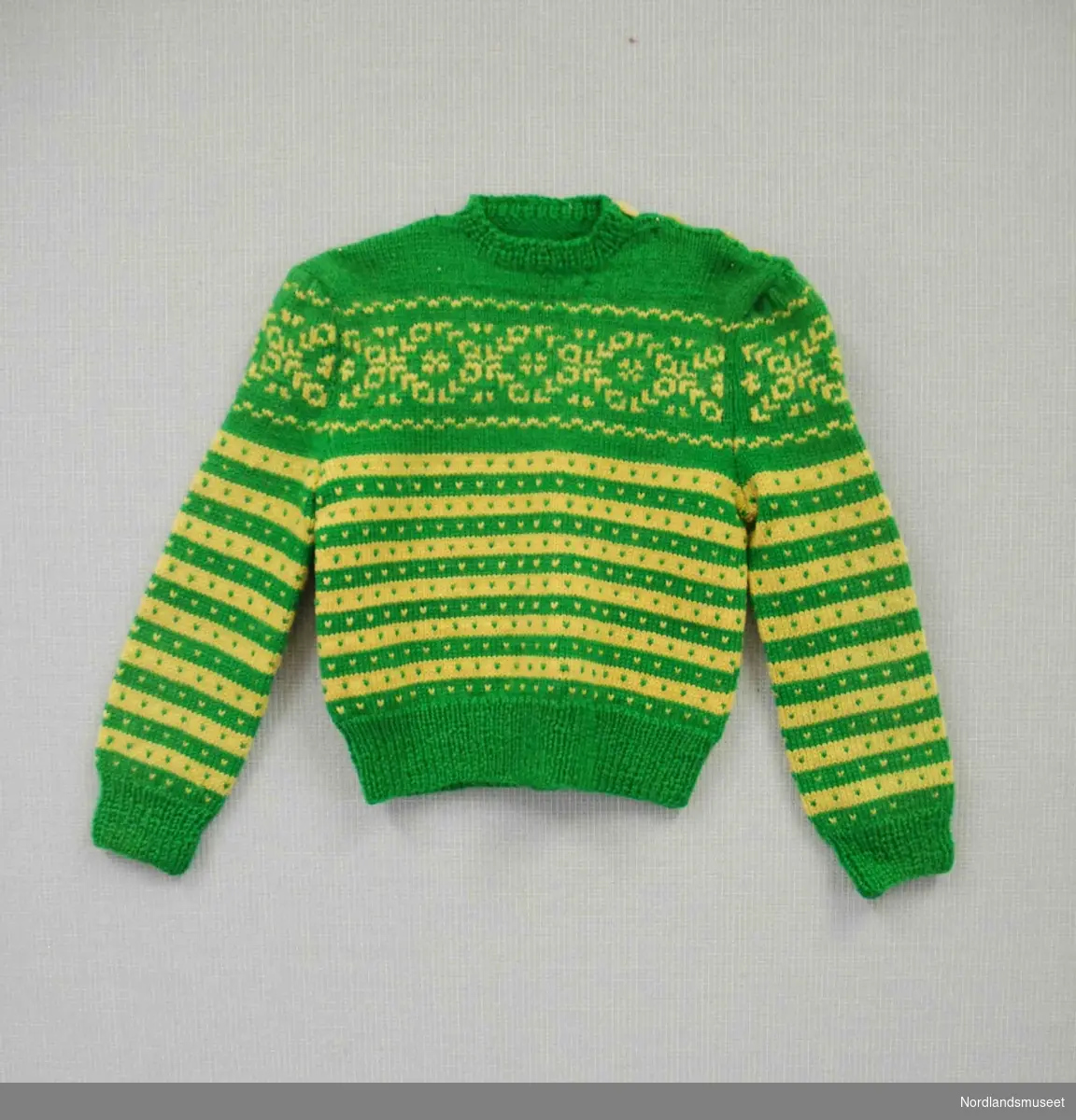 Grønn strikket genser med mønster i gult. Vrangbord i hals, på ermer og nederst. Åpning på venstre skulder med fire knapper til lukking.