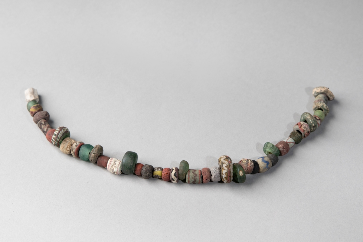 Pärlhalsband av glaspärlor samt 4 bronsspiralpärlor, vilka inte är fastsatta på halsbandet. Pärlorna är fasetterade, melon- och tunnformade och rikt ornerade i olika färger.