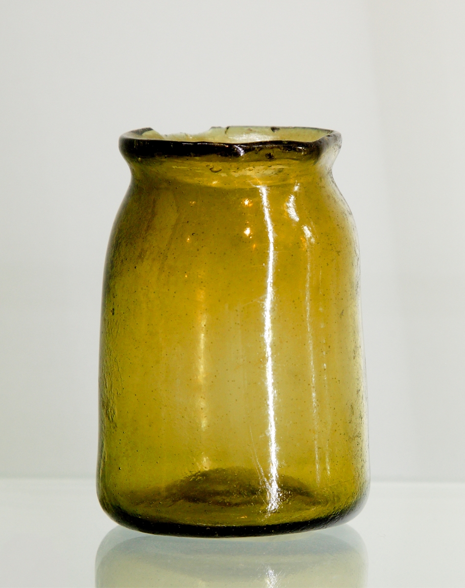 Apoteksglas i av grönbrun glasmassa, sk waldglas. Svagt konisk, avsmalnande sista del och utvikt mynning.