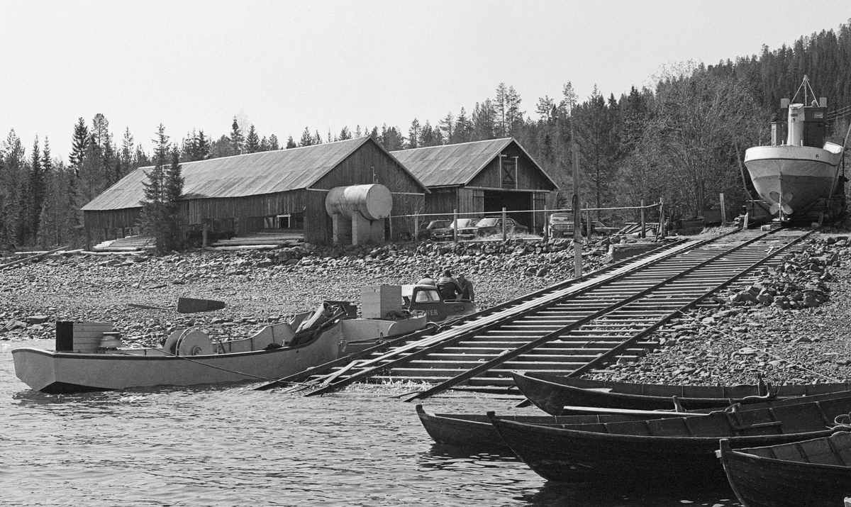 Fra Sørlistøa ved Osensjøen, som ligger i grensetraktene mellom Åmot og Trysil kommuner i Hedmark. Sjøen er om lag 47 kvadratkilometer stor og har et nedslagsfelt på om lag 1 300 kvadratkilomter. På cirka halvparten av dette arealet vokser det produktiv skog, så her var det mye fløtingsvirke. I 1939 kjøpte Glomma fellesfløtingsforening et jordfstykke på 21 dekar ved Sørlistøa, mellom strandsona på vestsida av innsjøen og bygdevegen. Her reiste de ei mannskaps- og kontorbrakke med tilhørende utedo, smie, et digert ved- og lensehus og et nesten lige stort båthus, og de bygde en slipp for sjøsetting og opptak av båter som ble brukt under fløtinga. Dette fotogreafiet er tatt våren 1984, idet lokale mannskaper forberedte det som skulle bli den siste fløtingssesongen her. Øverst på slippen ser vi akternende på den 55 fot lange og 12 fot brede slepebåten «Trysilknut», som på dette tidspunktet forlengst var ombygd fra damp- til dieseldrift. Bygningen til venstre er det 226 kvadratmeter store ved- og lensehuset, som etter at slepebåten hadde fått dieselmotor, først og fremst var et vinterlager for lensetømmer til bommene som ble brukt under bukseringa av tømmeret. Drivstoff til slepebåten og andre motordrevne farkoster ble på dette tidspunktet lagret i den digre ståltanken ved lensehusets nordvestre gavlvegg. Bak lensehuset ser vi båthuset, som hadde ei grunnflate på neten 200 kvadratmeter. Her oppbevarte Glomma fellesfløtingsforening inspeksjonsbåten «Ener», varpebåtene «Fløtarlars», «Lykkjeknut» og «Sjuluspær» samt 5-6 robåter. Disse farkostene ble sjøsatt og hentet tilbake til vinteropplagsstedet ved hjelp av ei vogn på en skinnegang som gikk mot vannet parallelt med, men tett inntil den som var bygd for Trysilknut. Da dette fotografiet ble tatt lå to av varpebåtene (Fløtarlars og Lykkjeknut) og inspeksjonsbåten, som alle hadde stålskrog, fortøyd til venstre for slippen, mens robåtene av tre lå til trutning til høyre i forgrunnen, i le av en molo som skulle skjerme innseilingsstedet mot de kraftige bølgene nordenvinden kunne utvikle på denne innsjøen.