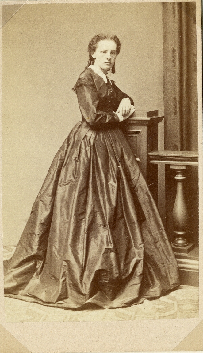 Porträttfoto av en kvinna i mörk sidenklänning och vit spetskrage med dito manschetter. Hon lutar sig mot en pelare vid en balustrad. I bakgrunden skymtar ett draperi. 
Helfigur, halvprofil. Ateljéfoto.