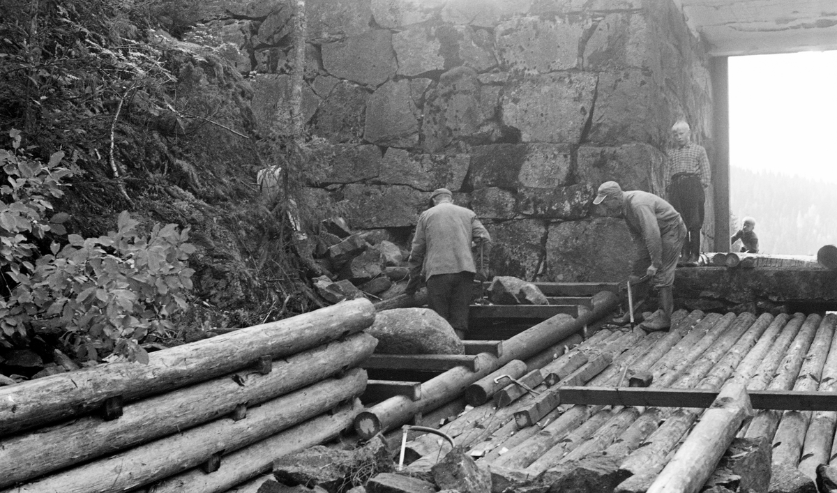 Reparasjon av ledeskjerm (skådam) ved damløpet i Måsåmyrdammen i Mørkåa, et vassdrag i Nord-Odal. Fotografiet ble tatt i 1956. På dette tidspunktet forsøkte man seg med kreosotimpregnerte materialer, i håp om at de skulle ha lang levetid i et fuktig miljø som dette. I bakgrunnen ser vi et høyt, tørrmurt damkar og en damåpning med golving av tømmer. Damløpet var tørrlagt da reparasjonsarbeidet ble utført. Da dette karet ble tatt var to voksne karer i gang med å bygge ei ny tømmerkiste som skulle fylles med stein inntil damkaret. To gutter fulgte arbeidet med interesse.