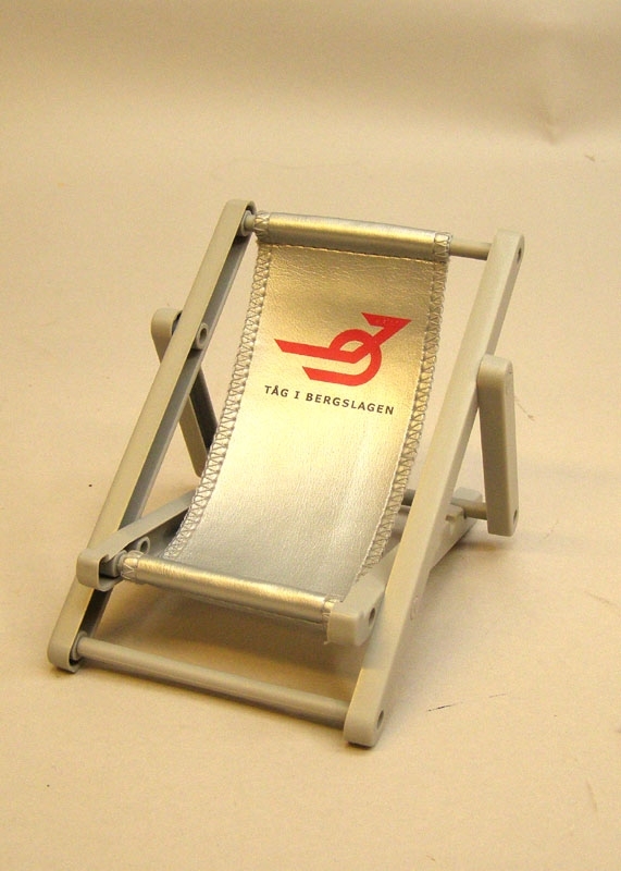 Bordsställ för mobiltelefon, i form av en solstol i miniatyr, grå ställning med ställbart läge.
Dynan är silverfärgad med röd logga för Tåg i Bergslagen.