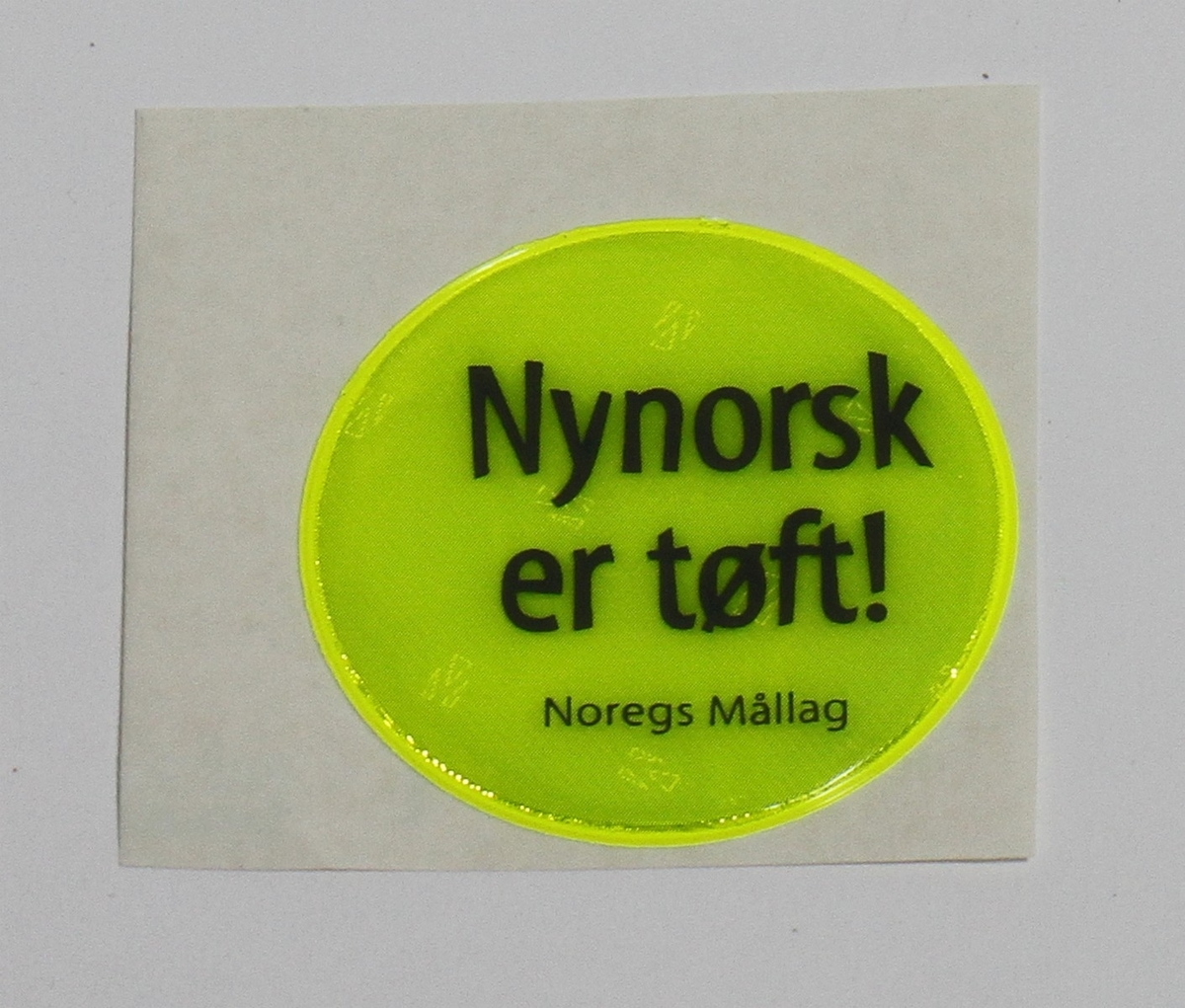 Gul klistrerefleks, som lyser i mørkret. Den sirkulære refleksen vart laga av Noregs Mållag, og bodskapen er at "Nynorsk er tøft!" Refleksen var truleg del av ein kampanje på 1990-talet.
