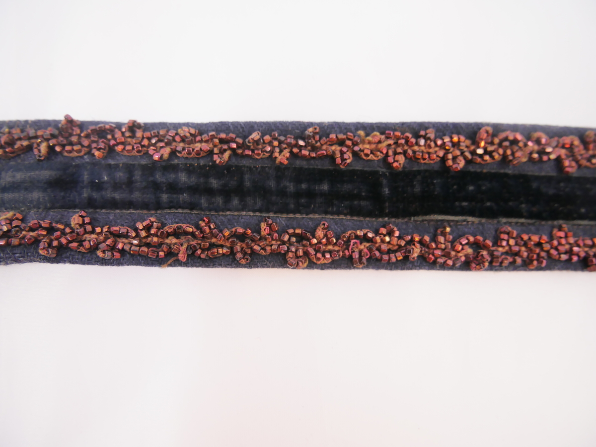 Mørkeblått belte med et svart fløyelsbånd midt på og to rader med brunrøde perler. En metallspenne med ankermotiv. 