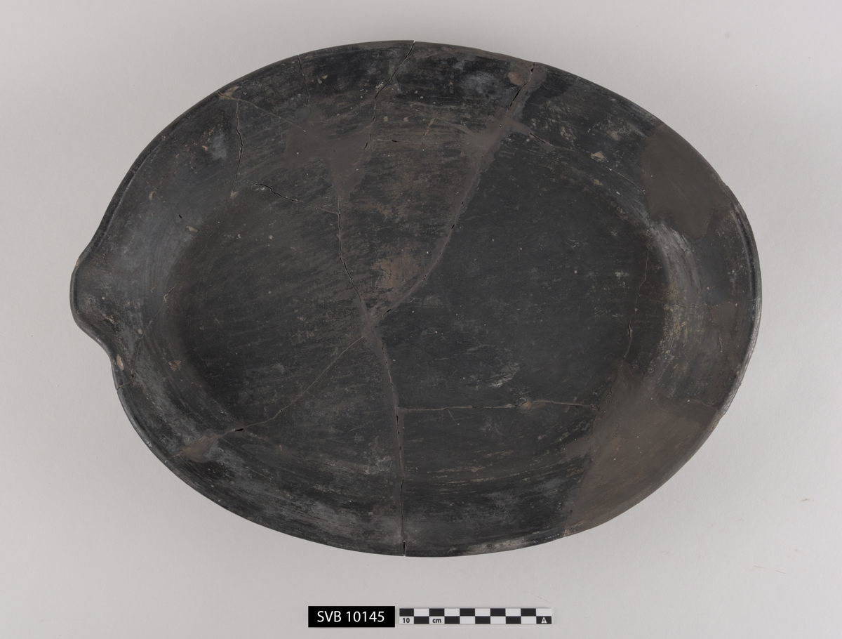 Ovalt kar av svartglasert keramikk med flat bunn og rett kant som er vinklet. Det er en helletut i den ene enden. Gjenstanden er sammensatt av originale og rekonstruerte deler.
