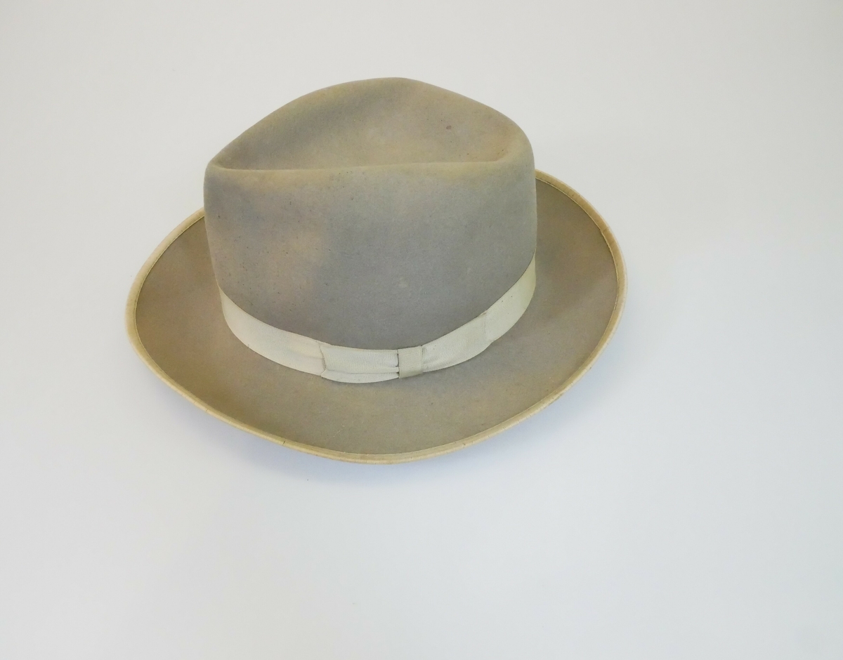 Grå herrehatt med pull og brem. Hvitt bånd med sløyfe rundt hattepullen og et hvitt bånd rundt bremen. Hvitt for med brun kant inne i hatten. 