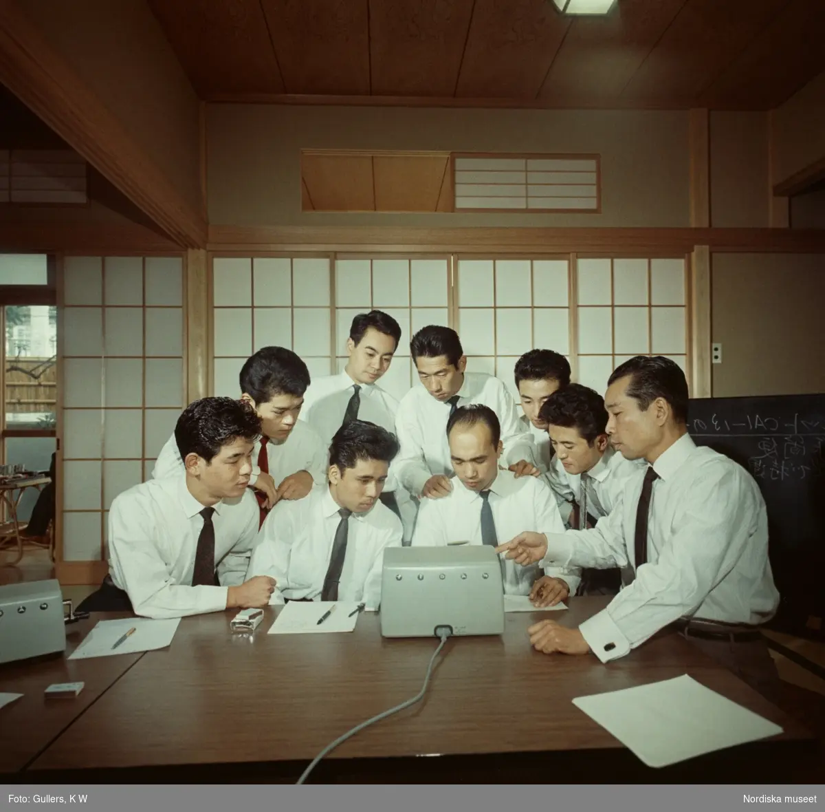 Facit. Nio män i män i vita skjortor och slips samlade runt en elektrisk kontorsmaskin.
