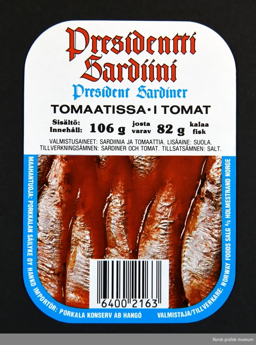 Etikett med hvit bakgrunnog detaljer i lys blå og rødt. Halve etiketten er dekt av bilde av innholdet i boksen: 
sardiner i tomat.
