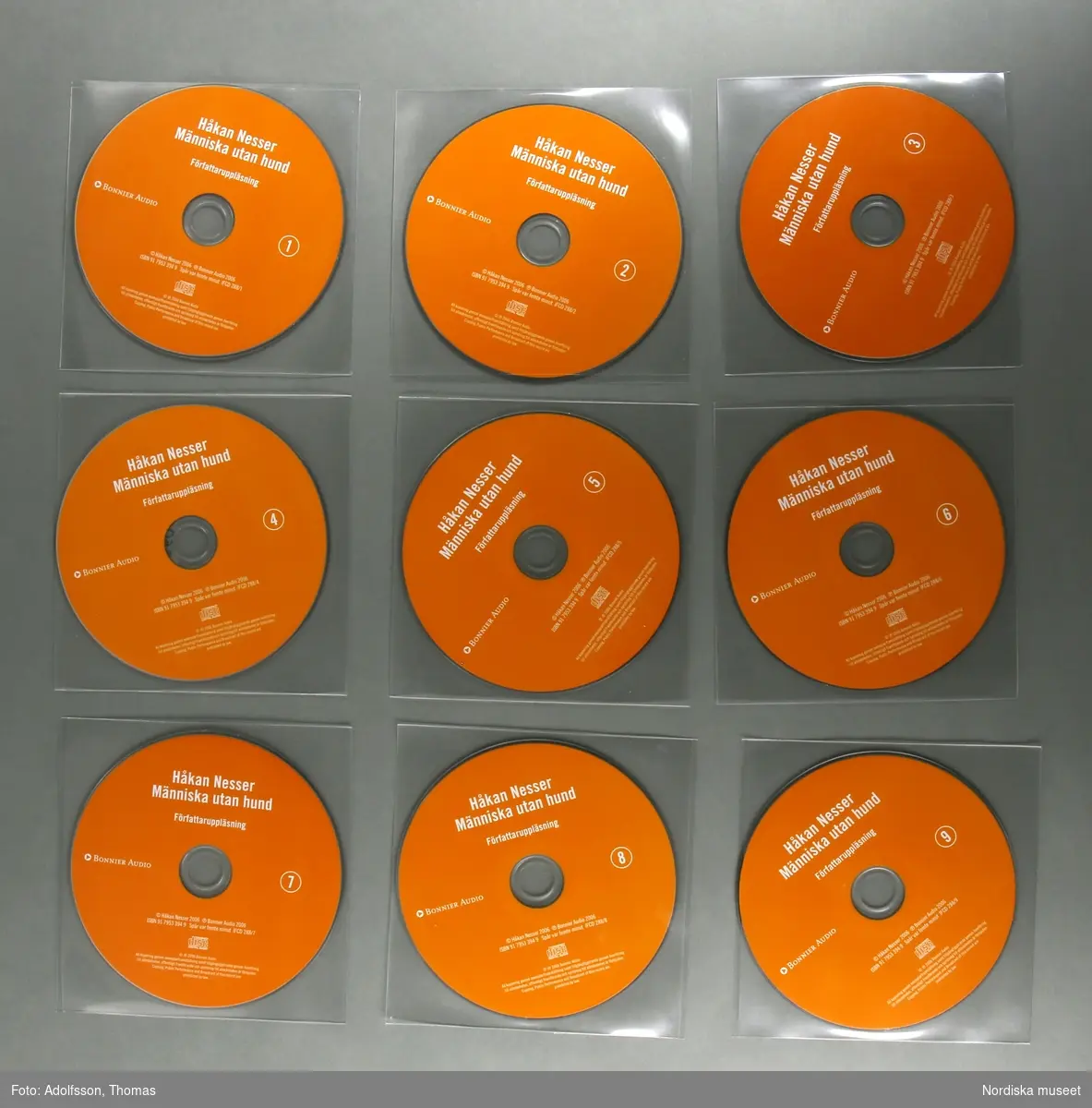 CD-bok.  a:1-2-n:1-2) 14 st CD-skivor, i en kvadratisk, transparent plastficka, innehållandes uppläsning av romanen "Människa utan hund" skriven av författaren Håkan Nesser som även är uppläsaren på denna CD-inspelning.  Inspelningen är totalt 15 timmar lång.

CD-skivorna är tunna cirkelfornade,  12 cm i diameter, på ena sidan oranga med text om titel samt kort om produktionen. Varje skiva är numrerad för att visa rangordningen i vilken skivorna ska lyssnas på/spelas. Baksidan på skivan är silverfärgad, speglliknande, och innehåller själva ljudspåren som läses av när skivan placeras i en CD-spelare.
Varje skiva är separat plackad i en kvadratisk, transparent, plastficka för att skydda mot repor som ev skulle kunna skada skivan. 
Samtliga skivor är tillsammans packade i en o) kvadratisk papperskartong/ konvolut. Konvolutet har på framsidan en bild av ett fönster med ett gultonat fotografi visandes ett fönster med en flaxande fågel utanför. På konvolutets baksida finns en introduktion till bokens handling samt  information om tillverkningen av denna CD-bok.
(CD är en förkortning av engelskans Compact Disc. CD-skivan kan lagra digital information, exempelvis musik.
CD skivans konstruktion: överst är ett transparant plastlager följt av ett tunt lager av blank metall med ett präglat spiralformat spår med mikroskopiskt små gropar som avläses av CD-spelarens laserstråle när skivan snurrar (ca 500-700 varv/minuten). Ljuset studsar tillbaka och läses som en följd av ettor och nollor, som i detta fall, blir till ljud.(Källa/. Nationalencyklopedin)


/Cecilia Wallquist 2019-02-20