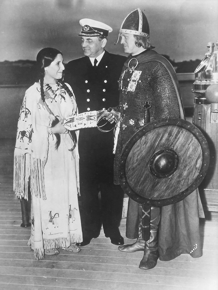 Ombord på Den Norske amerikalinje. En viking og en indianerkvinne overrekker hverandre gaver, kaptein Ingens står mellom.