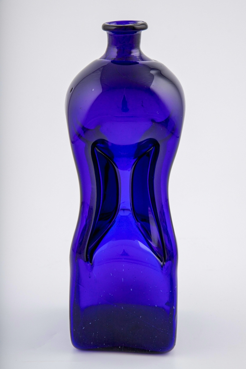 Høyreist klukkflaske i mørkt brungrønt glass. Timeglass-lignende utforming, hvor den kvadratiske underdelen er forbundet med overdelen gjennom et tynt rør. Lav hals med glatt kantring, samt puntemerke på undersiden.