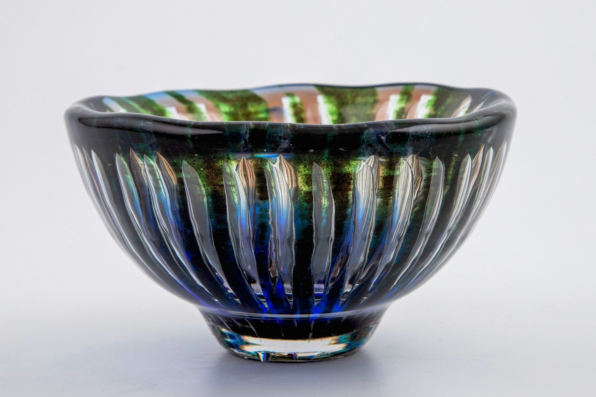 Ovalformet skål i "Ariel-glas", bestående av tykt gjennomskinnelig farget og klart glass. Grønne og beige striper stråler ut fra skålens bunn, hvorav førstnevnte er skapt ved hjelp av innsmeltet glitrende emaljefarge. Luftbobler mellom stripene. Et blåfarget parti er konsentrert på skålens nedre del av korpus samt fot.