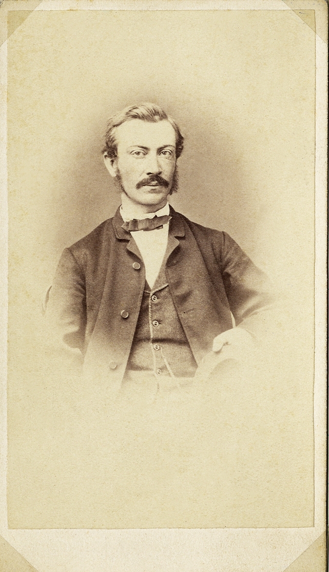 Foto av en man med mustascher, klädd i kavajkostym med väst, stärkkrage och fluga. På västen skymtar en klockkedja.
Midjebild, halvprofil, Ateljéfoto.