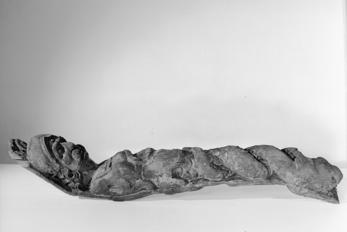 Triton med stora mustascher och lockigt hår. På huvudet sitter en hjälm med tudelad hjälmkam och nedhängande halsskydd i form av överlappande metallplåtar. Den rustningslika klädedräkten har en kvadratisk urringning. Kring midjan sitter ett band eller bälte. Den högra sidan av skulpturens bål upptas av en bladslinga som ersätter höger arm samt en del av midjebandet. Skulpturens fiskstjärt slingrar sig kring stjärten till en fisk eller en delfin vars huvud är synligt vid skulpturens fotända.

Skulpturens vänstra sida är inte skulpterad utan har utgjort en slät anliggningsyta som sträcker sig ned till det att fiskstjärten tar vid. Anliggningsytan är uppdelad på ett högre parti samt ett lägre part. Partierna är avgränsade genom ett vinkelformat hak i höjd med midjan. I det övre partiet återfinns en kvarsittande dymling. Bakstycket är kraftigt och snidat i ett med skulpturen. Skulpturens profil är svagt S-formad.
Skulpturen är relativt välbevarad.

Text in English: Triton with large moustache and long, curly hair. On the head he wears a smooth-surfaced helmet with upturned brim and hanging neck protector. A rudimentary crest can be seen on top of the helmet. The armour-like outfit has a square neck. There is a rudimentary girdle or belt round the waist. The arms are replaced by leafy ornamentation. Fastening surface opposite side left.

The fish tail is entwined with the tail of a fish or a dolphin which can be seen at the sculpture''s foot end. The figure is carved in one piece with a robust backboard. The profile is slightly S-shaped.
The sculpture is rather well preserved.