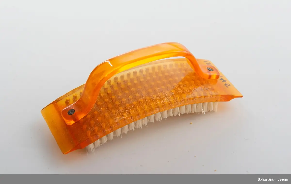 Konkav nagelborste i genomskinlig orange plast med bågformat handtag. 
Försedd med nylonborst.