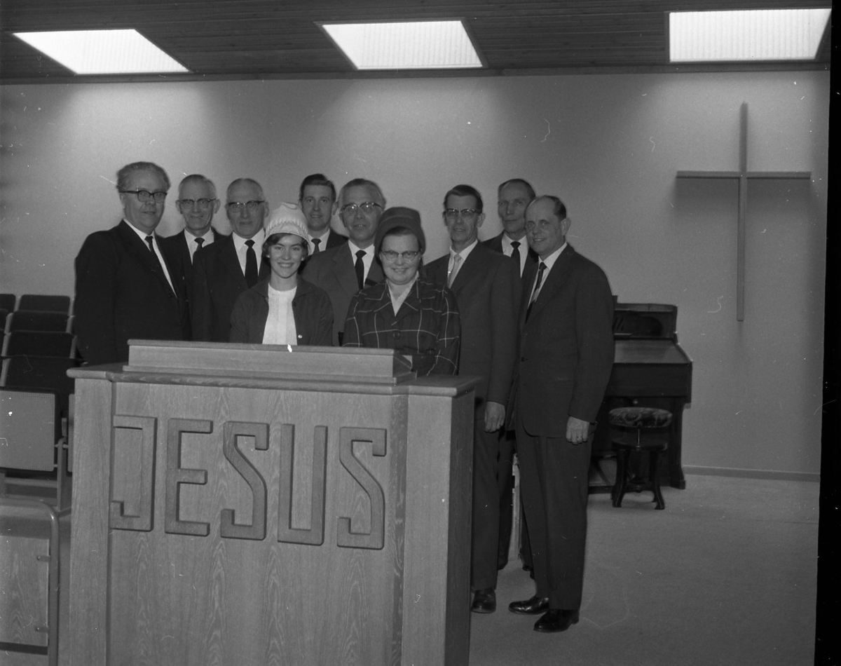 Åtta kostymklädda män och två kvinnor med hatt står bakom talarstolen i Filadelfiakyrkan. På densamma, mot salen, står texten "JESUS". Mannen som står mellan kvinnorna är pastor Robert Svensson.
