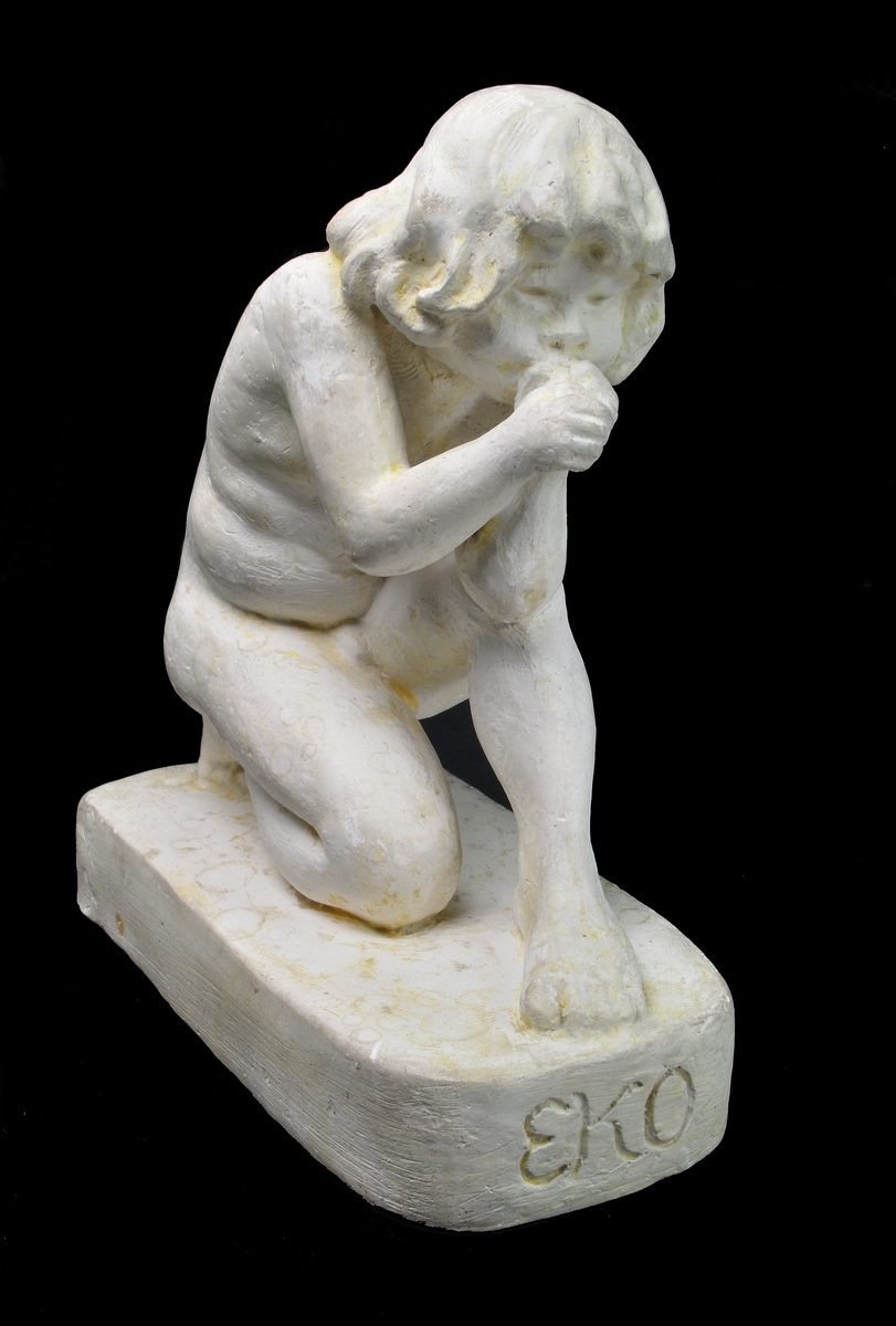 Skulptur i gips av Edith Norée: "Eko". Långhårig pojke sittande på huk med händerna framför munnen, formade för ljud. Rektangulär bottenplatta med inskription framtill: "EKO". Signatur baktill: "Paris Norée 1920".