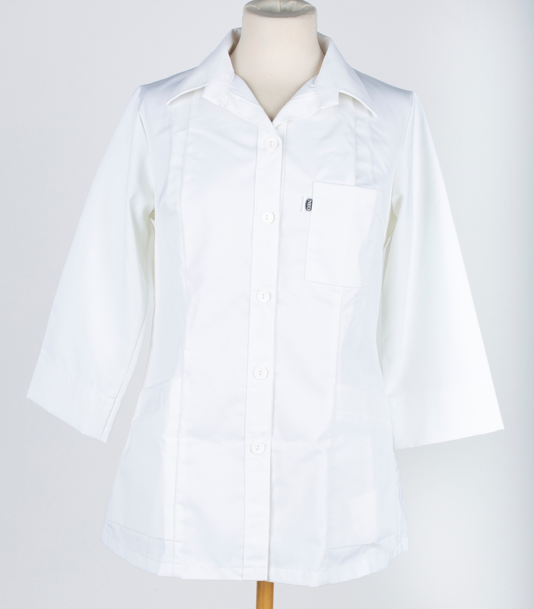 Skjorte, 7 bredder, med slag, 3 lommer, lange ermer, 3/4 lang.
Neo yrkesklær. Sannsynligvis laboratoriefrakk.