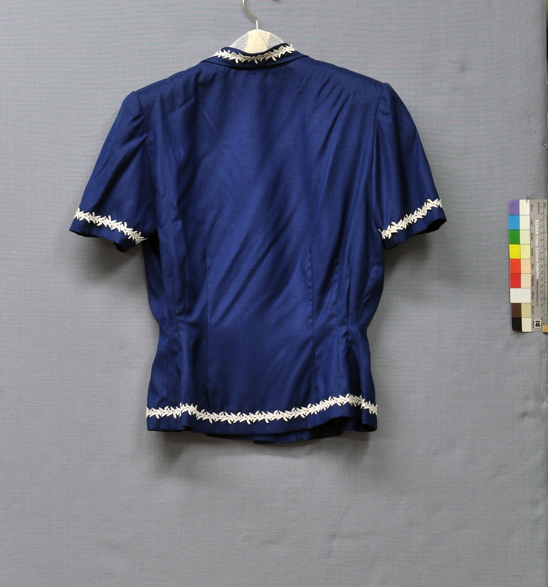 Mörkblå dräkt bestående av kortärmad jacka (BM63831:1) och kjol (BM63831:2) i silke och viskos. Jackans framsida delad, knäppt med 3 vita pärlemorknappar. 2 bystinsnitt, 4 midjeinsnitt. Baksidan helskuren med 6 midjeinsnitt. Schalkrage med snibbar. Kort ärm. Axelvaddar. Kragen, ärmarna och nederkanten garnerade med vit maskingjord blombård av bomull. 
Kjolen av 4 våder, rynkad. 25 mm bred linning knäppt med 2 hakar. Kjolens nederkant med samma blombård som på jackan. Blixtlås i vänster sida. Storlek 42.