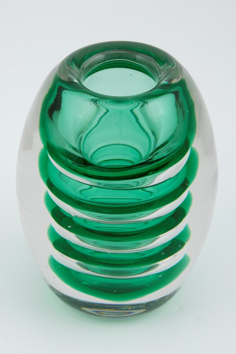 Ovalformet vase i underfangsglass. Et indre sjikt i grønt gjennomskinnelig glass, som er utformet som ringformede vulster, omgitt av et tykt lag av klart glass. Sirkulær åpning og planslipt bunn.