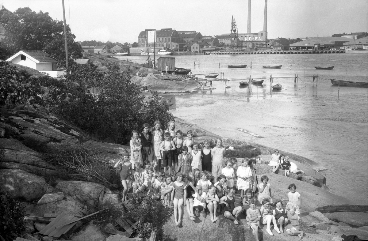 Badestrand på Kråkerøy i Fredrikstad med Denofa Fabrikker i bakgrunnen, antakelig. Barn og voksne i forgrunnen. .