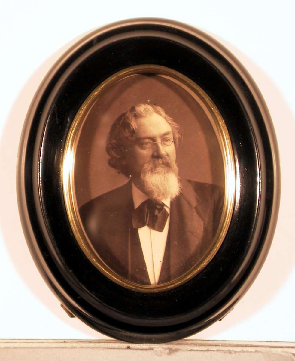 Portrettfotografi av en voksen mann med briller, halvlangt bølget hår og pent klippet skegg, ikledd dress med hvit skjorte og mørkt halsbind. Han ser skrått til venstre.