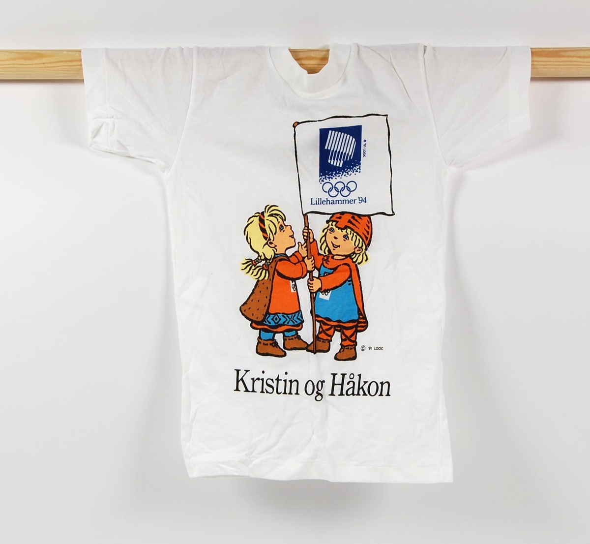 Hvit t-skjorte med flerfarget motiv av maskotene for de olympiske vinterleker på Lillehammer i 1994, Kristin og Håkon. Kristin og Håkon holder et flagg med logo fro de olympiske vinterleker på Lillehammer i 1994. T-skjorten er i barnestørrelse.